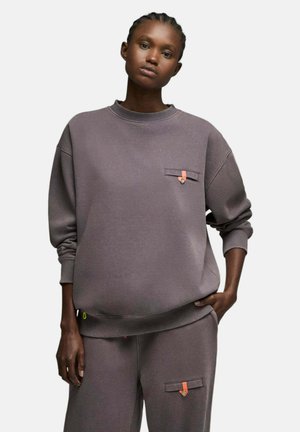 Femme portant un sweat-shirt ample gris foncé et un pantalon assorti avec de petits détails orange et des boutons sur un fond uni.