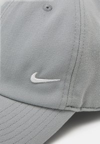 Szary kapelusz wykonany z teksturowanej tkaniny z zakrzywionym daszkiem. Na froncie znajduje się białe haftowane logo Nike. Widoczne są szwy wzdłuż krawędzi.