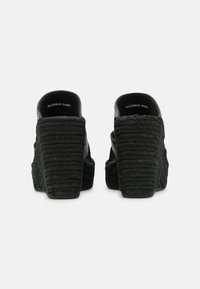 Svarta vävda kilklackar visade bakifrån med stängda hälar och texturerade plattformssulor på en vit bakgrund.