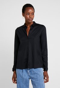 Schwarzes Langarmshirt mit Knopfleiste, Kragen und seitlichen Einschnitten. Hergestellt aus glattem Stoff, mit figurnaher Passform und klassischem Design.