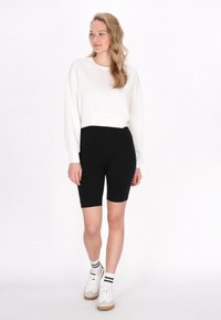 Bílá croptop mikina spárovaná s černými cyklistickými šortkami, s hladkou texturou. Modelka má na sobě bílé tenisky s černými pruhovanými ponožkami.