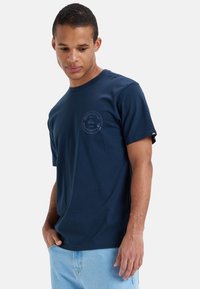Camiseta de algodón azul marino con cuello redondo, que presenta un diseño de logo circular en el lado izquierdo del pecho y mangas cortas. Textura suave.