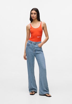 Punane soonikkerega bodi valge äärisega, kombineeritud kõrge vöökohaga helesiniste laiade pükstega ja mustade varbavahe plätude paariga, ühevärvilise tausta ees.