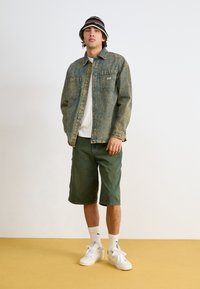 Giubbotto di jeans azzurro chiaro con due tasche sul petto, t-shirt bianca, pantaloni corti verdi e sneakers bianche, sopra un cappello a secchiello a strisce bianche e nere.