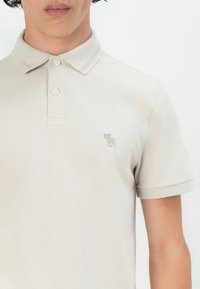 Polo shirt crem cu un material texturat, cu un plastron cu trei nasturi și un mic logo verde brodat pe pieptul stâng. Mâneci scurte.