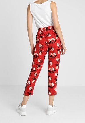 Vrouw draagt rode hoge taille broek met witte bloemenprint, gecombineerd met een witte mouwloze top en witte sneakers, staand tegen een effen achtergrond.