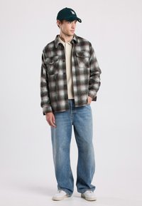 Geruite flanellen overhemd in groen en bruin met twee borstzakken, gecombineerd met een beige polo en lichtblauwe brede jeans, witte sneakers.