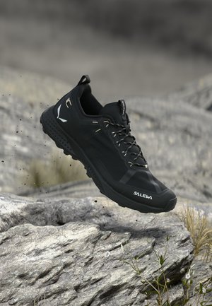Chaussure de trail running Salewa noire en l'air au-dessus d'un terrain rocheux avec de petites plantes, mettant en avant l'adhérence et l'utilisation en extérieur.