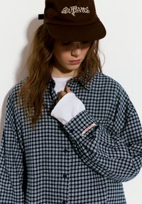 Camicia di flanella a quadri blu navy e bianca, con design a bottoni e un piccolo accento rosso sulla manica. Maglietta bianca visibile sotto.