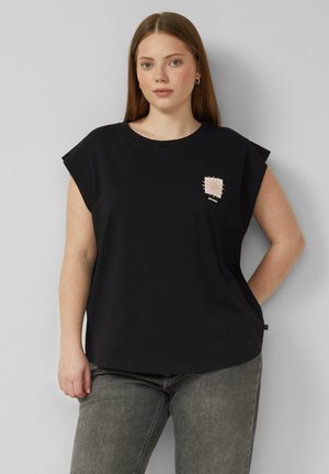 Junge Frau mit glatten braunen Haaren trägt ein schwarzes ärmelloses Shirt mit kleinem Sonnenmotiv und ausgewaschenen grauen Jeans, stehend vor einer grauen Wand.