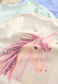 T-shirt couleur crème avec un graphique de licorne, une crinière en sequins roses et des accents scintillants. Deux t-shirts bleu clair sont visibles en arrière-plan.