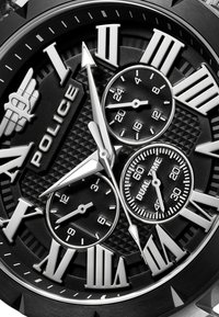 Melna metāla pulksteņa ar romiešu cipariem, fakturēta melna pulksteņa saskarne, sudraba akcenti un vairāki subpulksteņi, kas attēlo atšķirīgas laika funkcijas.