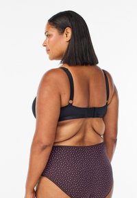 Žena s vlasmi po pleciach, oblečená v čiernej podprsenke a vysokých fialových nohavičkách so vzorom, zobrazená zozadu na bielom pozadí.