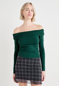 Grønn off-shoulder langermet topp i myk stoff, med rynkede detaljer ved halsen, kombinert med et svart rutete skjørt.