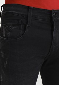 Los jeans de mezclilla negra presentan un ajuste slim, un ligero desgaste y un diseño clásico de cinco bolsillos. La cintura incluye un pasador para cinturón y un cierre de botón.