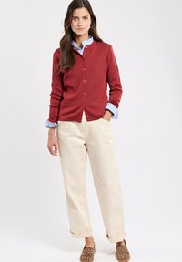 Cardigan rosso con colletto a coste e tasche, abbinato a polsini di camicia azzurri chiari in vista. Pantaloni beige arrotolati e scarpe con stampa leopardata.