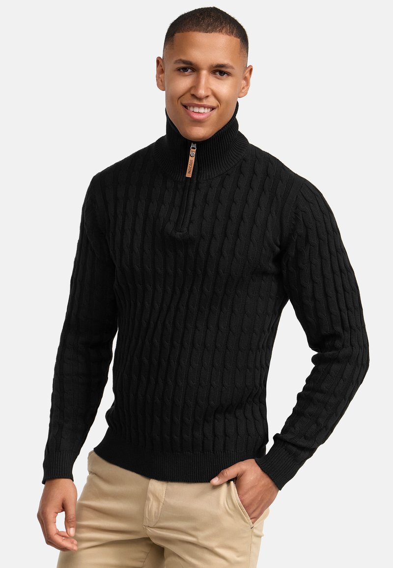 Schwarzer Strickpullover mit strukturiertem Zopfmuster, hohem Kragen und halbem Reißverschluss. Gerippte Bündchen und Saum. Das Modell trägt hellfarbene Hosen.