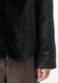 Sort tekstureret jakke med faux fur kant og guld "M" logo på ærmeåbningen, båret med mørkebrune læderbukser, hånd afslappet ved siden.