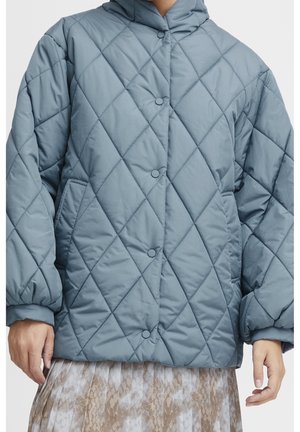 Veste matelassée bleue avec boutons-pression, poches latérales et poignets élastiqués, portée sur une jupe beige à motifs.