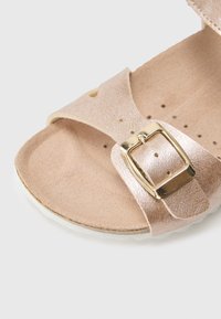 Rózsaarany papucs texturált pánttal, arany csattal, puha talpbéléssel és légáteresztő lyukakkal. Vékony, fehér talp.