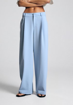 Person iført lyseblå, vidde bukser med høj talje og sorte sandaler med åbne tæer, stående med krydsede arme foran en ensfarvet baggrund.