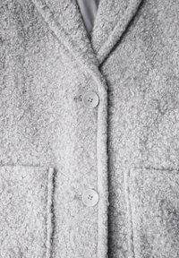 Grå texturerad kappa i boucletyg, med två framfickor och vita knappar. Kragen och slag är distinkt utformade.