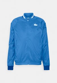 Blå lätt bomberjacka med hel dragkedja, ribbade ärmar och nederkant, samt en vit krage med en liten broderad logotyp på bröstet.