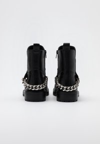 Botas negras hasta el tobillo con parte superior de cuero suave, tacón bajo, cremallera en la parte trasera y detalle de cadena metálica en la parte posterior para mayor textura.