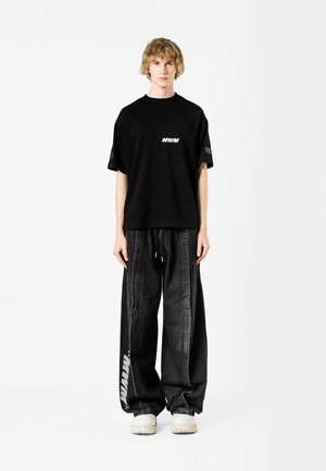 Camiseta negra con el logo blanco "MWM", combinada con pantalones de mezclilla negros holgados que presentan un diseño de franjas laterales y una cintura elástica.
