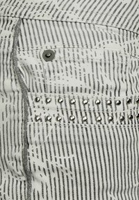 Weißer Denim-Stoff mit schwarzen vertikalen Streifen. Verfügt über einen kleinen Metallknopf und metallische Nieten entlang der Gesäßtasche zur Dekoration.