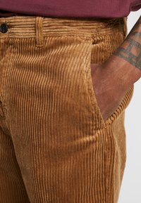 Brązowe spodnie sztruksowe z pionowym prążkowaniem, wyposażone w boczne kieszenie, z zapięciem na guzik i teksturowaną powierzchnią.