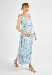 Rochie de maternitate florală de culoare albastră deschisă, cu bretele voluminoase, talie strânsă și tiv cu volane. Asortată cu sandale aurii cu barete.
