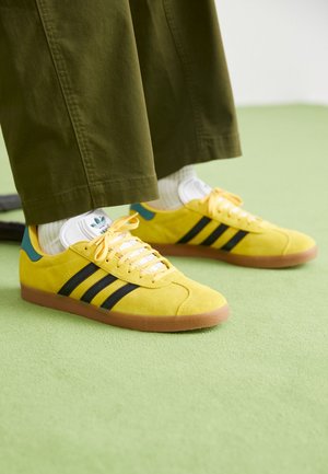 Żółte buty sportowe Adidas z czarnymi paskami, noszone z białymi skarpetkami i oliwkowo zielonymi spodniami, stojące na jasnozielonej podłodze.