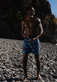 Zwemshorts in marineblauw met een levendig palmbomenpatroon, gemaakt van lichtgewicht stof. Model staat op een kiezelstrand, blootsvoets.