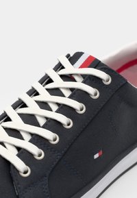 Marine canvas sneakers met witte veters, metalen oogjes en subtiele rood-blauw-witte accenten op de tong; rubberen zool en textiele textuur.