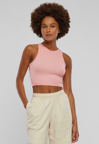 Vaaleanpunainen, hihaton crop-top, joka on valmistettu ribbitetystä kankaasta. Topissa on pyöreä pääntie ja tiukka malli, joka on yhdistetty vaaleanbeigeihin, löysiin shortseihin.