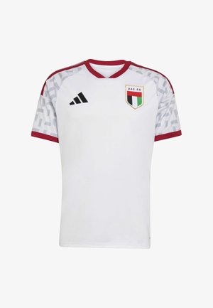 Biała koszulka piłkarska z czerwonymi akcentami, krótkie rękawy, dekolt w serek, wzorzyste ramiona oraz herb Zjednoczonych Emiratów Arabskich (UAE FA) po lewej stronie piersi. Logo Adidas po prawej stronie.