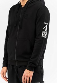 Crna zip-up dukserica od mekanog materijala, s prednjim džepom, kapuljačom na vezanje i bijelim tekstualnim detaljima na lijevoj ruci.