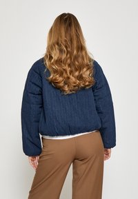 Zilās denim pufjaka ar īsu dizainu, ribinātu apakšmalu un nedaudz par lielu izmēru; satur dūrienu paneļus un teksturētu virsmu.