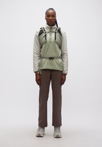 Γυναίκα που φοράει ανοιχτό πράσινο φλις πουλόβερ, γκρι γιλέκο πεζοπορίας, καφέ παντελόνι και γκρι μποτάκια πεζοπορίας, στέκεται μπροστά σε απλό λευκό φόντο.