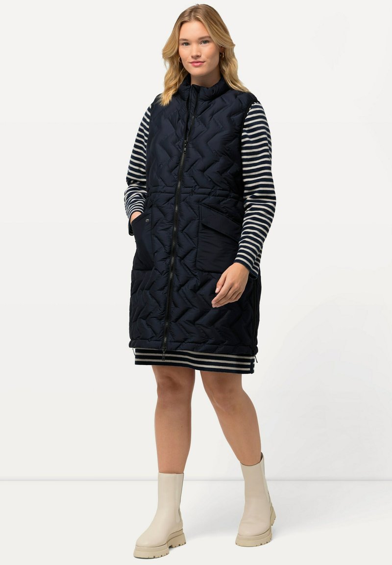 Navy gesteppte ärmellose Weste mit Reißverschluss, vorderen Taschen und Chevron-Muster. Tragen über einem schwarz-weiß gestreiften Langarmkleid. Beige Knöchelschuhe.