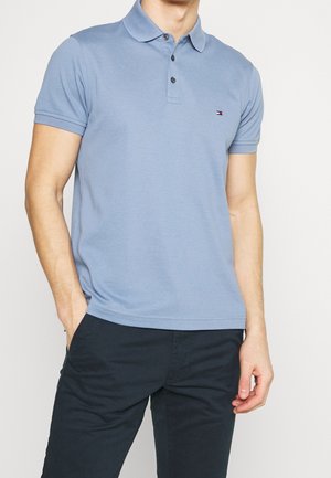 Mann trägt ein hellblaues kurzärmeliges Poloshirt mit kleinem Logo auf der Brust und dunkle Hose, eine Hand in der Tasche.