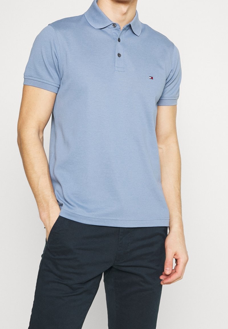 Homme portant un polo bleu clair à manches courtes avec un petit logo sur la poitrine et un pantalon foncé, une main dans la poche.