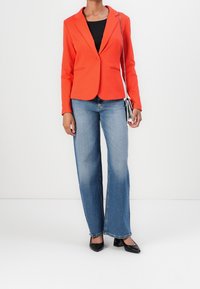 Blazer entallado rojo con solapas de muesca, cierre de un solo botón y dos bolsillos frontales, combinado con jeans de pierna ancha en azul claro y tacones negros.