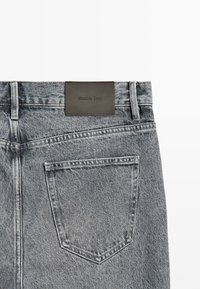 Zadní pohled na vybledlé šedé džínové kalhoty se zobrazující jedním kapsou a černou obdélníkovou značkou přišitou na pasu.