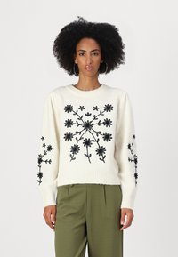 Maglione crema con ricamo floreale nero, scollatura rotonda a coste e maniche lunghe. Abbinato a pantaloni verde oliva.