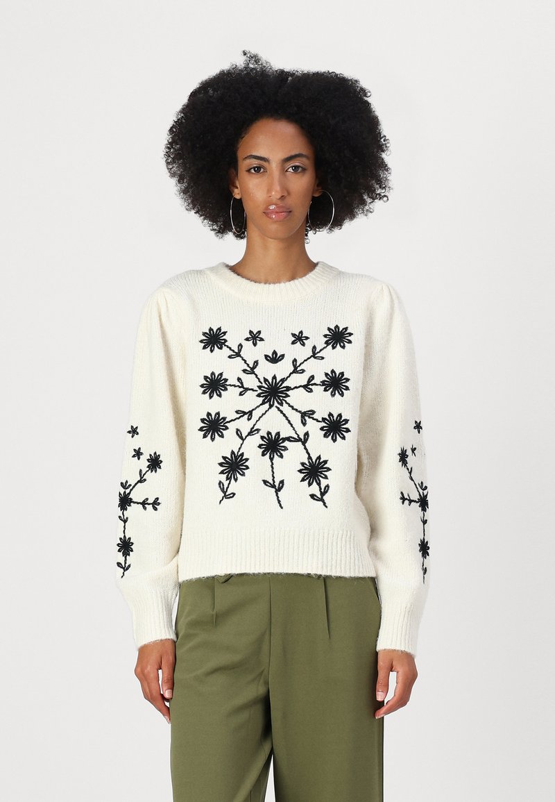 Maglione crema con ricamo floreale nero, scollatura rotonda a coste e maniche lunghe. Abbinato a pantaloni verde oliva.