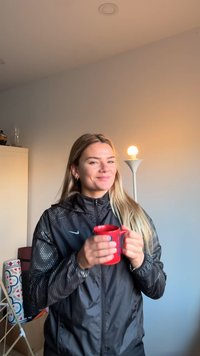 Rødt krus med håndtak holdt foran en person iført en svart jakke med teksturerte ermer, stående ved en lampe med en glødende pære.