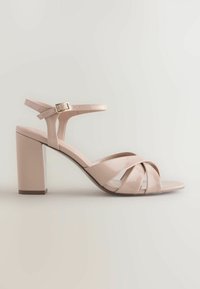 Sandale à talon haut en cuir verni beige avec talon bloc, brides croisées sur le devant et bride de cheville avec boucle dorée.