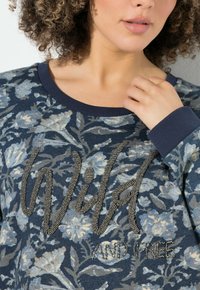 Mujer con cabello rizado vestida con un suéter floral azul que tiene un texto de cuentas que dice "Salvaje y Libre" tocando el cuello cerca del collar.